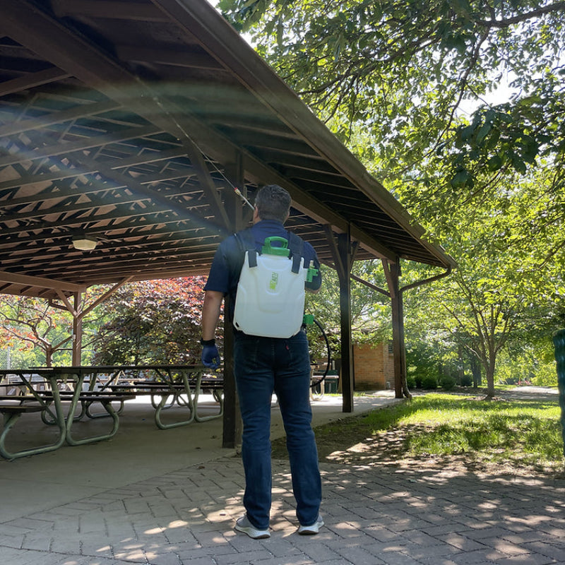 Rear outdoor view of a person wearing a FlowZone Storm 2.5 10L battery-powered backpack sprayer, spraying under a wooden pavilion roof. The white tank with green lid and black straps is clearly visible, highlighting portability and hands-free spraying convenience.