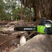 Greenworks 40V Brushless Chainsaw resting on a large fallen log in a wooded forest area. The green and black tool with visible chain bar is surrounded by bark, leaves, and trees, emphasizing durability and field-ready performance in outdoor conditions.