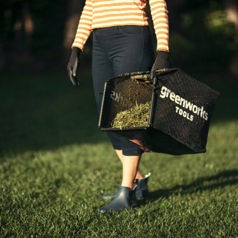 Person carrying a Greenworks Tools grass catcher filled with clippings across a freshly cut lawn. The black catcher with white logo is held beside dark pants and boots, in warm sunlight with a soft-focus green garden background.