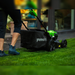 Person mowing grass with the Greenworks 40V 46cm Push Mower, showing its black grass catcher with white logo and bright green body. Scene set in a backyard with lush green lawn, deck area, and modern house in the background.