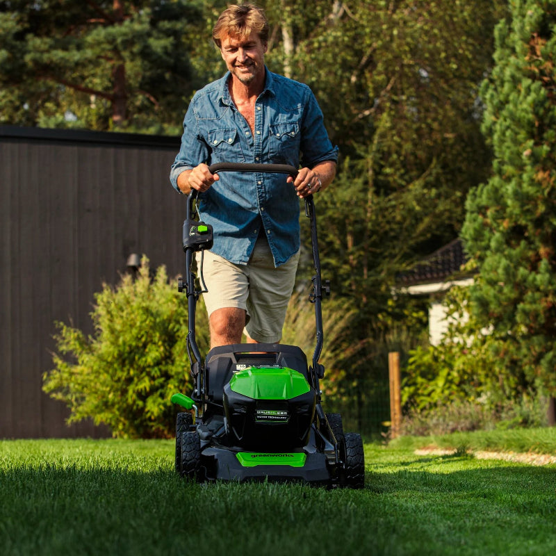 Person mowing a lush green lawn with the Greenworks 40V 46cm Push Mower. The bright green mower is seen from the front with black wheels and handle, set in a sunny backyard surrounded by trees and garden foliage.