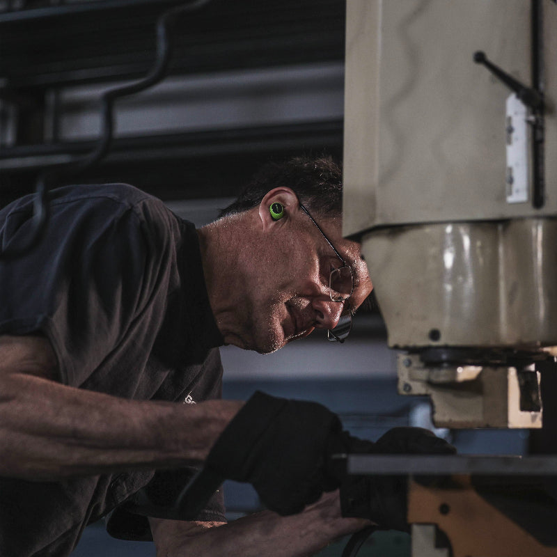 ISOtunes Free 2.0 wireless earbuds worn by a tradesperson operating industrial machinery, providing Bluetooth audio and in-ear hearing protection to reduce noise exposure while allowing communication during workshop and manufacturing tasks.