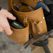 Worker placing ISOtunes Free Aware Bluetooth earbuds case into a leather tool belt pocket, showing durable design and portability for jobsite hearing protection.