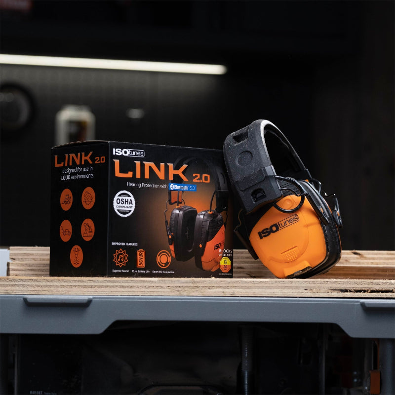 ISOtunes Link 2.0 Bluetooth hearing protection earmuffs shown beside retail box on a workshop bench, featuring over-ear noise reduction and wireless audio, designed for use in loud work environments such as construction, fabrication, and machinery operation.