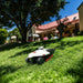 A Mammotion LUBA AWD X robotic lawn mower operates on a sloped, sunlit lawn surrounded by trees and a large house with a red roof in the background, under a clear blue sky.