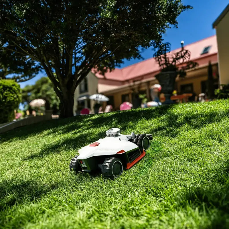 A Mammotion LUBA AWD X robotic lawn mower operates on a sloped, sunlit lawn surrounded by trees and a large house with a red roof in the background, under a clear blue sky.