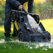 A person rinses the underside of a Mammotion LUBA Mini robotic lawn mower with a hose, showing water splashing on its wheels and blades. The mower is positioned on wet green grass with a bright, blurred background.