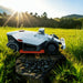 A Mammotion LUBA AWD X robotic lawn mower sits on rocky terrain surrounded by tall green grass, with morning sunlight streaming through trees and distant hills in the background.