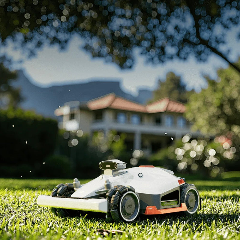 Mammotion Luba AWD robotic lawn mower cutting grass under a shaded tree, with a large house and mountains blurred in the background, demonstrating its ability to mow in shaded garden areas with precision and ease.