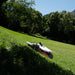 Mammotion Luba AWD X robotic mower cutting grass on a steep, sunlit slope. Surrounded by lush greenery and tall trees, the mower demonstrates its ability to handle uneven, slanted terrain while maintaining a neat, trimmed lawn.