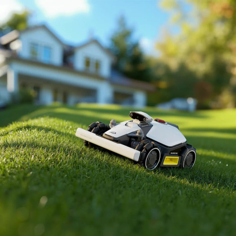 Mammotion Luba Mini AWD robotic mower cutting grass on a sloped lawn in front of a modern suburban house, with sunlight illuminating the green yard and trees in the softly blurred background.
