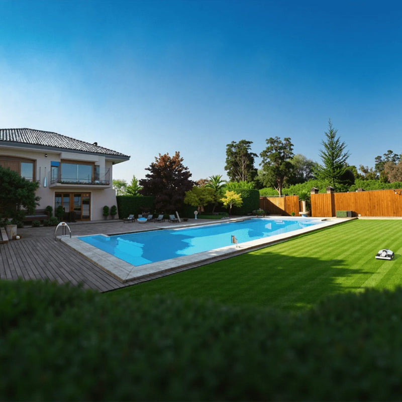 A Mammotion Luba Mini robotic lawn mower trims a neatly striped green lawn beside a bright blue swimming pool, surrounded by tall hedges, trees, and a modern two-story house with a wooden deck under a clear blue sky.