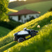 A Mammotion Luba Mini robotic lawn mower with a white and black body and yellow trim climbs a steep grassy hill surrounded by lush greenery, trees, and a blurred house with a red roof in the background under warm sunlight.