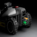 Close-up of the Mammotion Luba Mini robotic lawn mower showing its rear section with large rugged wheels and a removable black battery featuring orange latches and a glowing green arrow indicator, set against a dark background.
