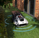 A Mammotion YUKA robotic mower operates on grass beside a brick house wall and hedge, with blue sensor waves illustrating its smart navigation and obstacle detection under soft daylight.