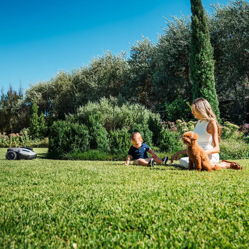 A woman and child sit on a sunny green lawn with a small dog, while the Mammotion YUKA robotic mower works nearby, showcasing safe and quiet operation in a peaceful garden surrounded by tall trees and bushes.