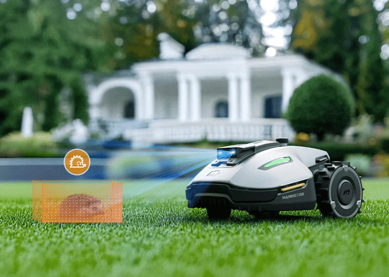 A Mammotion Yuka Mini robotic lawn mower with spiked wheels and green lights detects a hedgehog inside an orange safety box on a green lawn, with a large white house and trimmed bushes visible in the background