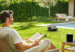 A man sits on a patio chair reading a book beside a table with a black speaker, while a Mammotion Yuka Mini robotic lawn mower operates on green grass near a swimming pool, with lounge chairs and tall hedges in the background.