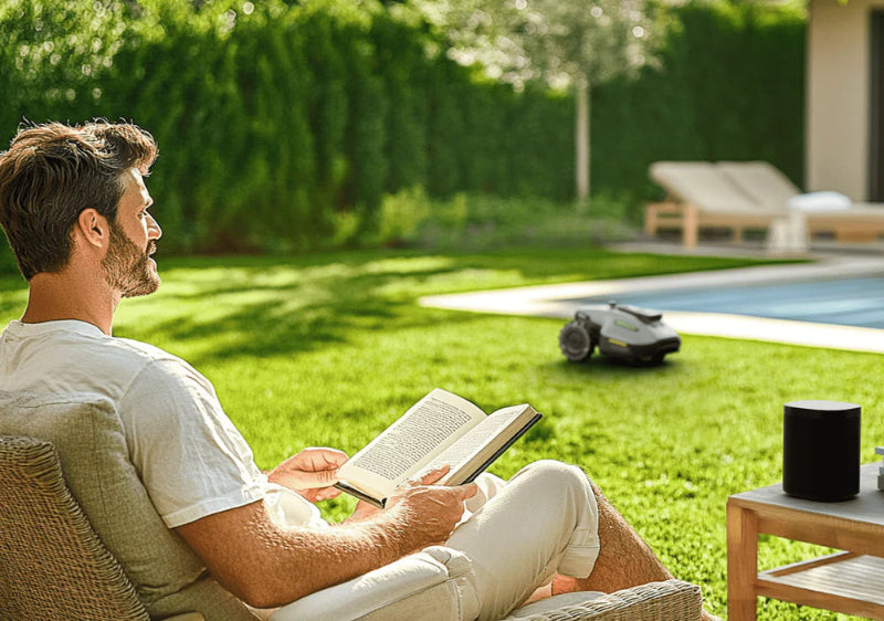 A man sits on a patio chair reading a book beside a table with a black speaker, while a Mammotion Yuka Mini robotic lawn mower operates on green grass near a swimming pool, with lounge chairs and tall hedges in the background.