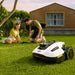 A white Mammotion Yuka Mini robotic lawn mower operates on green grass while a man and young girl plant a tree nearby, surrounded by potted flowers, wooden planters, and a brick house with large windows in the background.