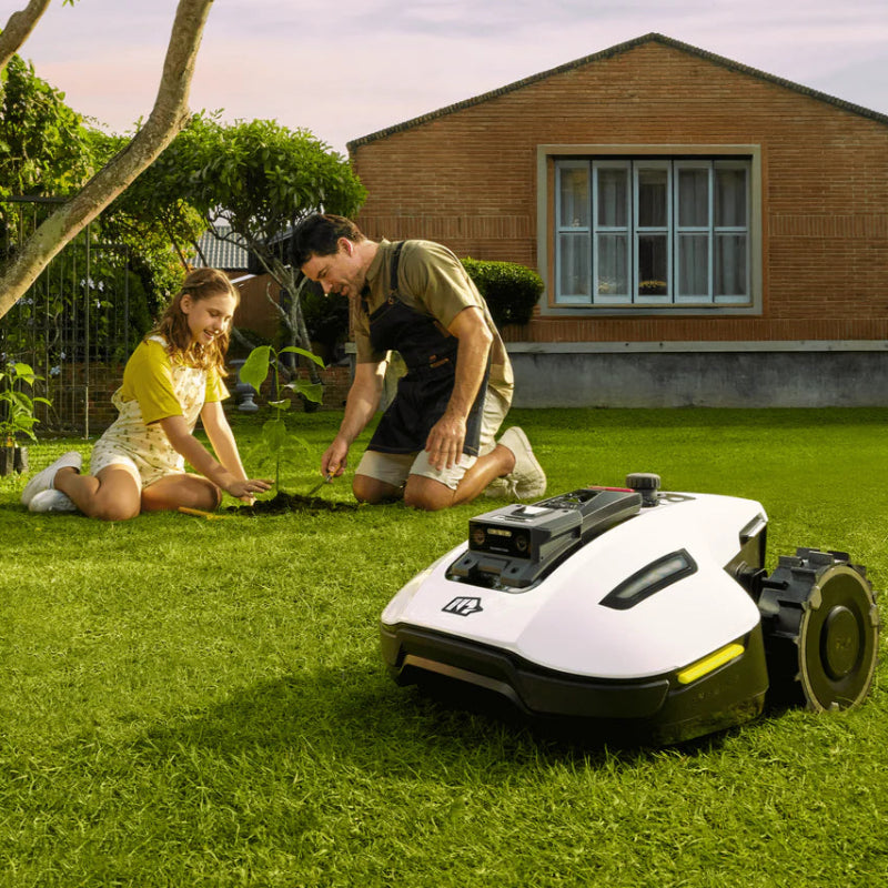 A white Mammotion Yuka Mini robotic lawn mower operates on green grass while a man and young girl plant a tree nearby, surrounded by potted flowers, wooden planters, and a brick house with large windows in the background.