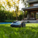 A Mammotion Yuka Mini robotic lawn mower in gray with rugged spiked wheels and glowing green side lights, positioned on freshly cut green grass. In the background, a modern house with a wooden deck, outdoor sofa, and potted plants is visible, surrounded by blurred leafy trees in bright daylight.