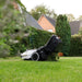 Mammotion Yuka robotic mower operating on a residential lawn, collecting freshly cut grass into its rear-mounted collection bag, with houses, hedges, and tall trees in the background.