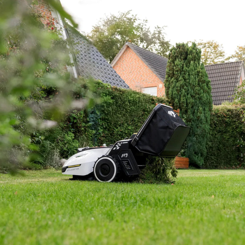 Mammotion Yuka robotic mower operating on a residential lawn, collecting freshly cut grass into its rear-mounted collection bag, with houses, hedges, and tall trees in the background.