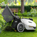 Mammotion Yuka robotic mower with attached lawn sweeper kit shown collecting clippings on a lush green lawn, surrounded by bushes and flowers in a landscaped garden.