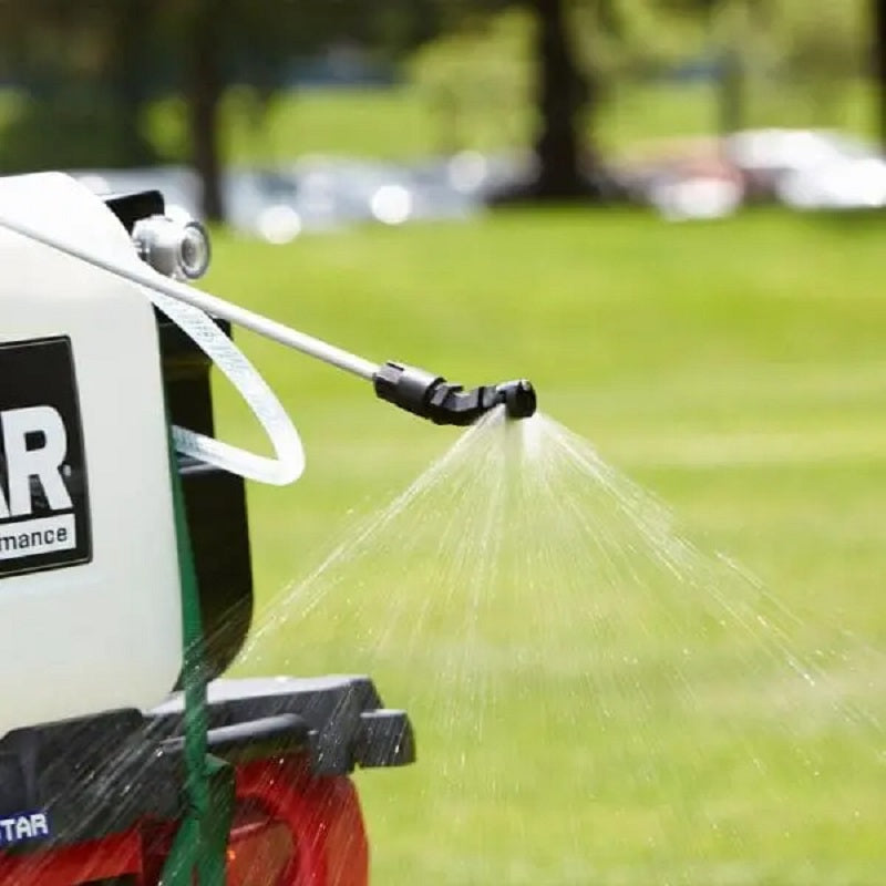 NorthStar 12V ATV sprayer spraying water evenly across green grass. The image shows the nozzle in action, demonstrating fine mist coverage from the mounted white tank for efficient lawn, garden, or agricultural spraying.