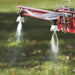 Close-up of NorthStar tow-behind sprayer boom showing dual nozzles spraying fine mist over green grass. Features red metal frame, clear tubing, and even spray pattern in outdoor sunlight for precision lawn and garden treatment.