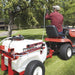 NorthStar tow-behind boom and spot sprayer in use, attached to a red garden tractor. The operator sprays trees on a green lawn using the hose system. Features a white tank, red frame, and durable pneumatic tires.