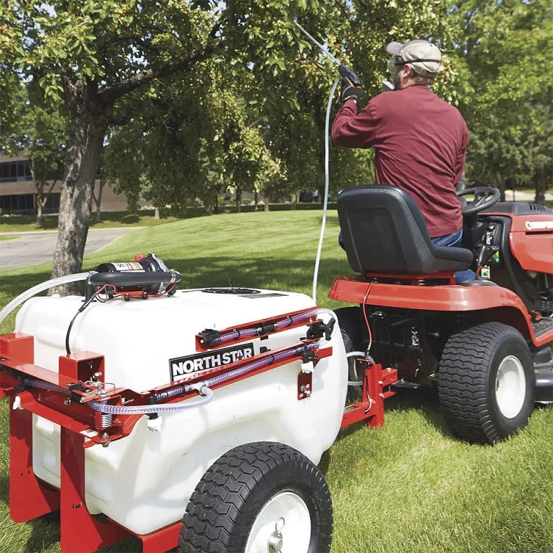 NorthStar tow-behind boom and spot sprayer in use, attached to a red garden tractor. The operator sprays trees on a green lawn using the hose system. Features a white tank, red frame, and durable pneumatic tires.