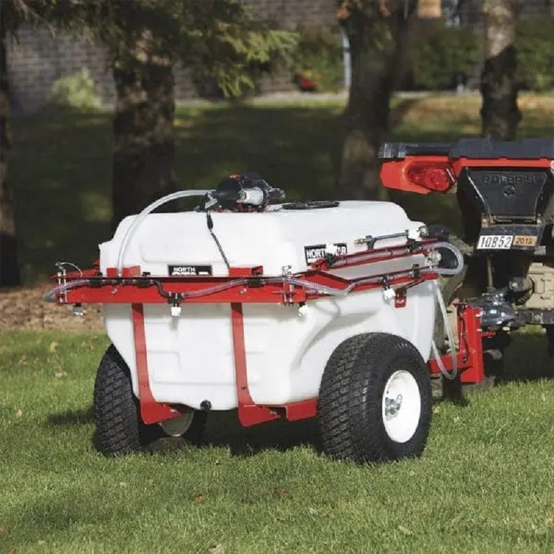 NorthStar 383-litre tow-behind boom and spot sprayer attached to an ATV, spraying on green lawn. Features a large white tank, red frame, durable tires, and mounted pump system, shown in an outdoor garden setting for lawn care application.