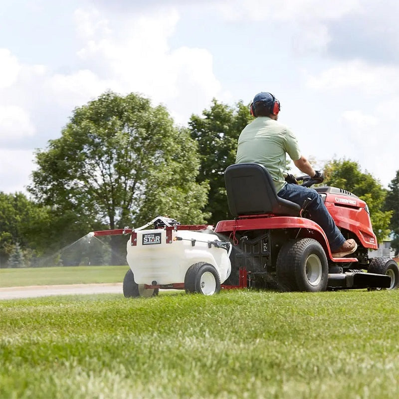 NorthStar tow-behind boom and spot sprayer attached to a red lawn tractor spraying green grass. Features a white tank, red frame, and wide wheels. Shown outdoors on a bright sunny day with trees in the background.