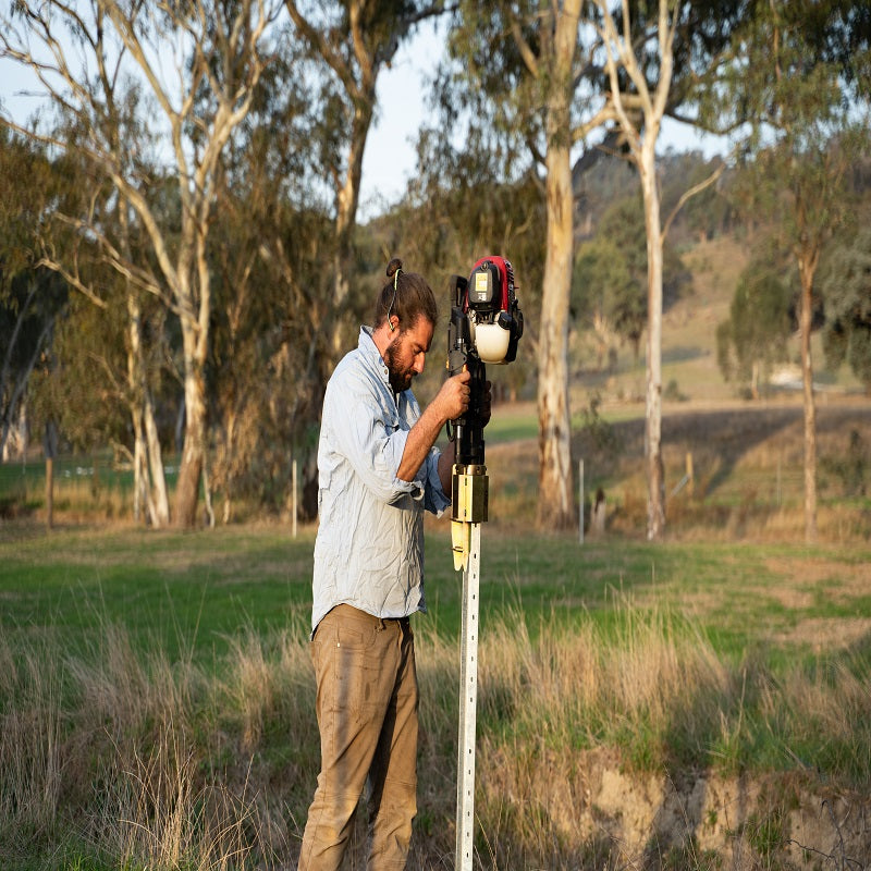 Worker carrying a Powertech PPD-80 post driver powered by a Honda GX engine, walking through a paddock with a mallet in hand and a utility vehicle in the background.