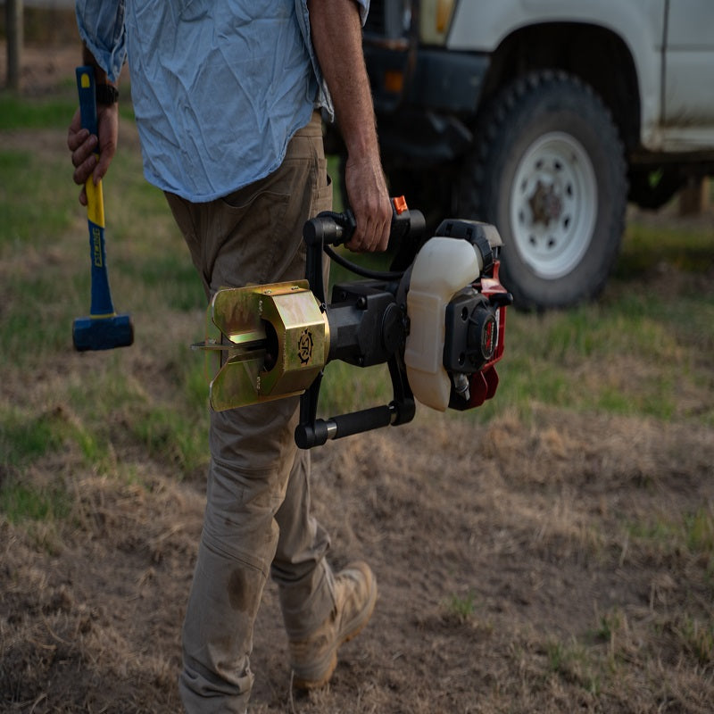 Worker carrying a Powertech PPD-80 post driver powered by a Honda GX engine, walking through a paddock with a mallet in hand and a utility vehicle in the background.
