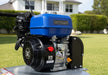 Close-up of the Predator LS460 lawn scarifier engine featuring the blue GB225 motor with black housing, recoil starter, and metal frame. Photographed outdoors on a bright day with a house and green lawn in the background.