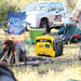 Rato R2500iS yellow inverter generator in use at a campsite, placed near a campfire with flames burning, surrounded by camping gear including a chair, table, cooler, and a parked vehicle in the background under daylight conditions.